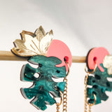 Tropicana Earrings: Colorful, handcrafted earrings shaped like green monstera leaves with gold leaf details and pink circular tops, hanging from a gold chain on a rod against a light background for a tropical-inspired design.