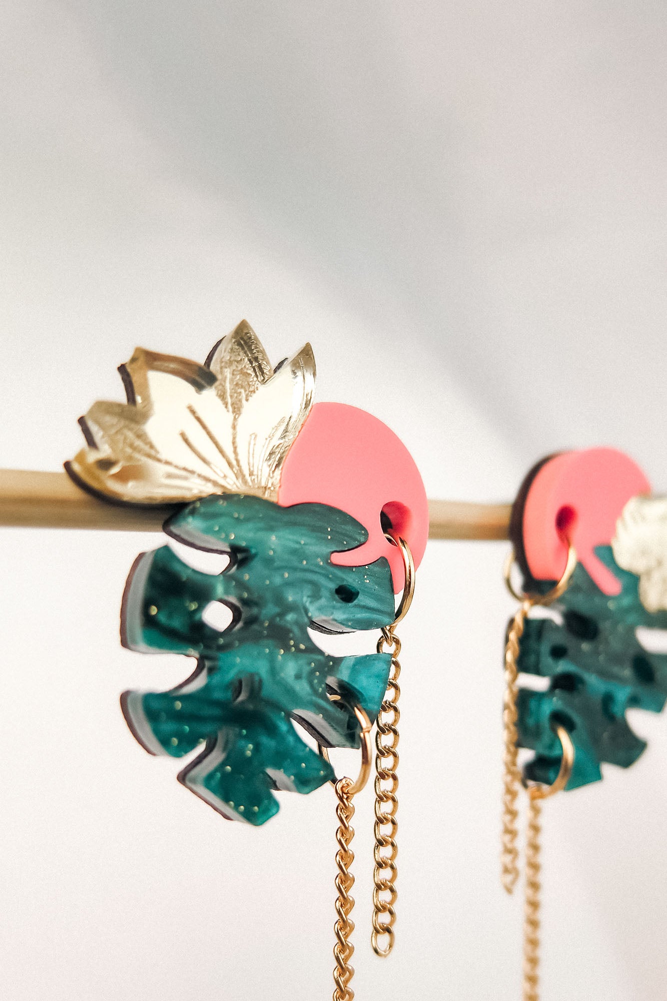 Tropicana Earrings: Colorful, handcrafted earrings shaped like green monstera leaves with gold leaf details and pink circular tops, hanging from a gold chain on a rod against a light background for a tropical-inspired design.