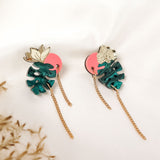 A pair of handcrafted Tropicana Earrings featuring green monstera leaves, gold floral accents, pink circular backings, and gold chain tassels, displayed on a white fabric background with dried grasses in the corner.