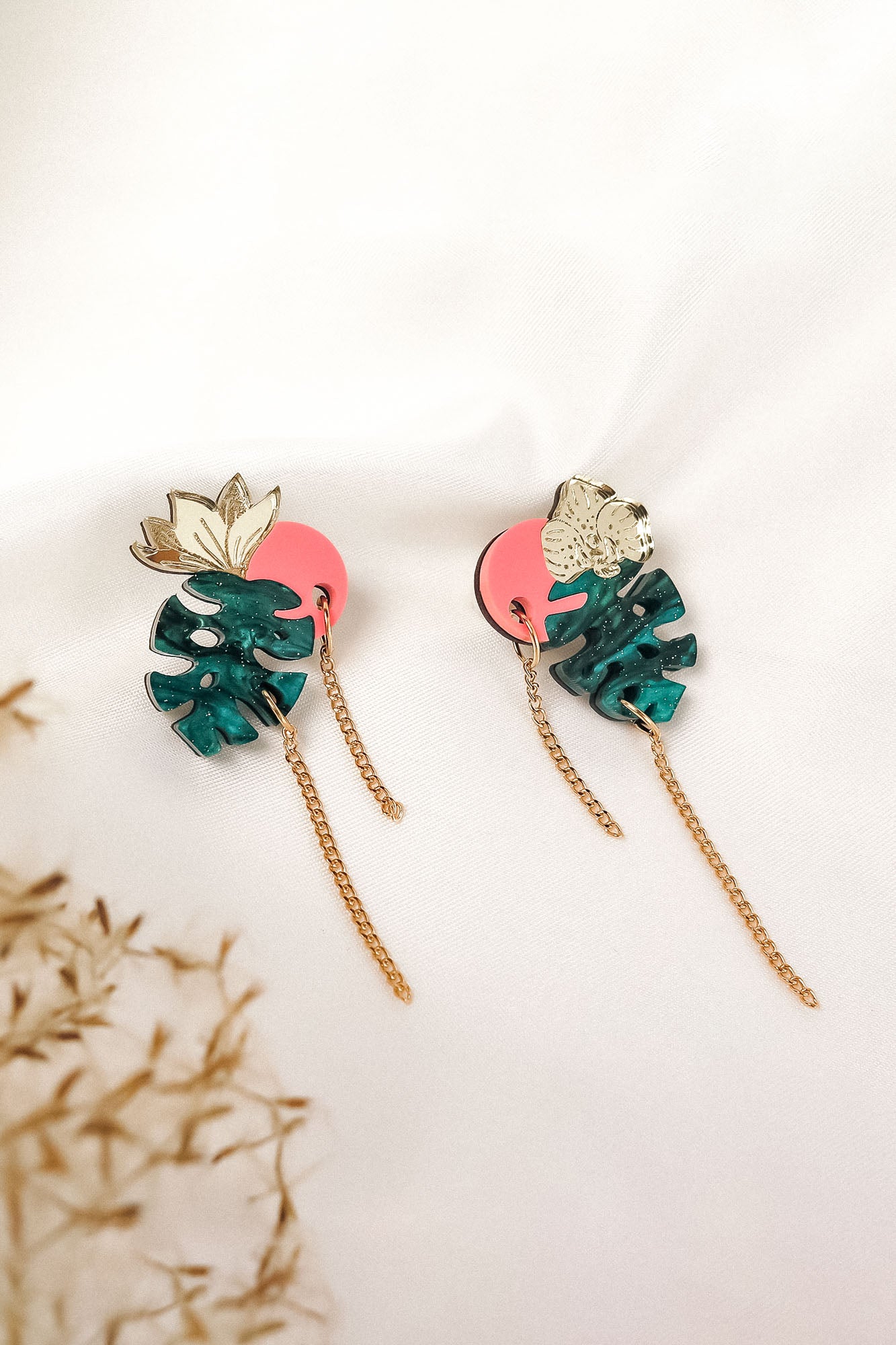 A pair of handcrafted Tropicana Earrings featuring green monstera leaves, gold floral accents, pink circular backings, and gold chain tassels, displayed on a white fabric background with dried grasses in the corner.