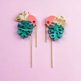 A pair of colorful Tropicana Earrings with gold leaves, teal monstera-shaped pendants, pink circular accents, and long gold chains, featuring a handcrafted, tropical-inspired design on a light purple background.