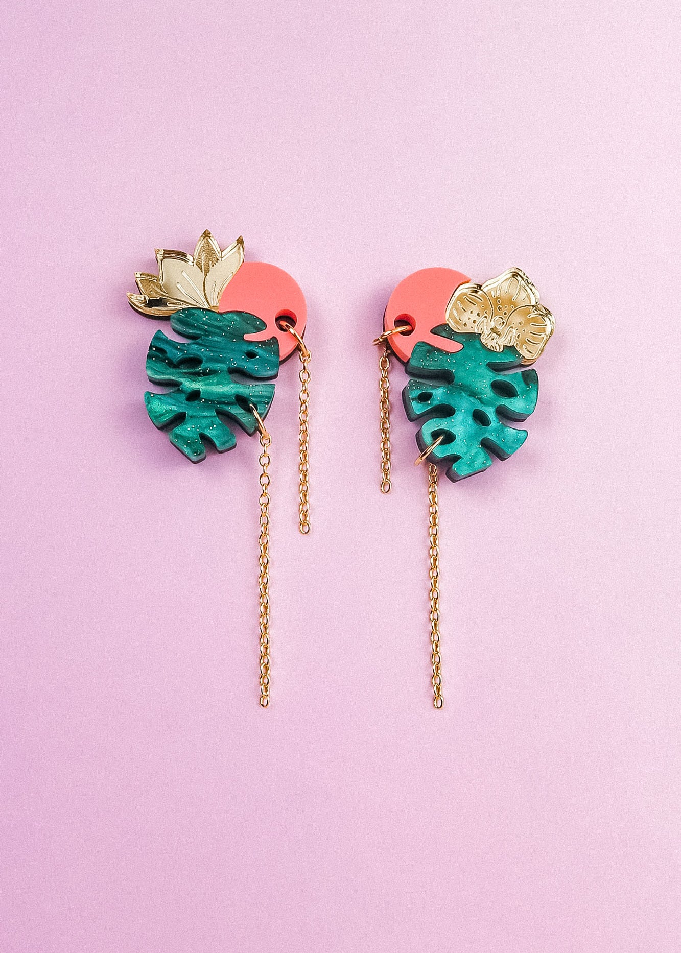A pair of colorful Tropicana Earrings with gold leaves, teal monstera-shaped pendants, pink circular accents, and long gold chains, featuring a handcrafted, tropical-inspired design on a light purple background.