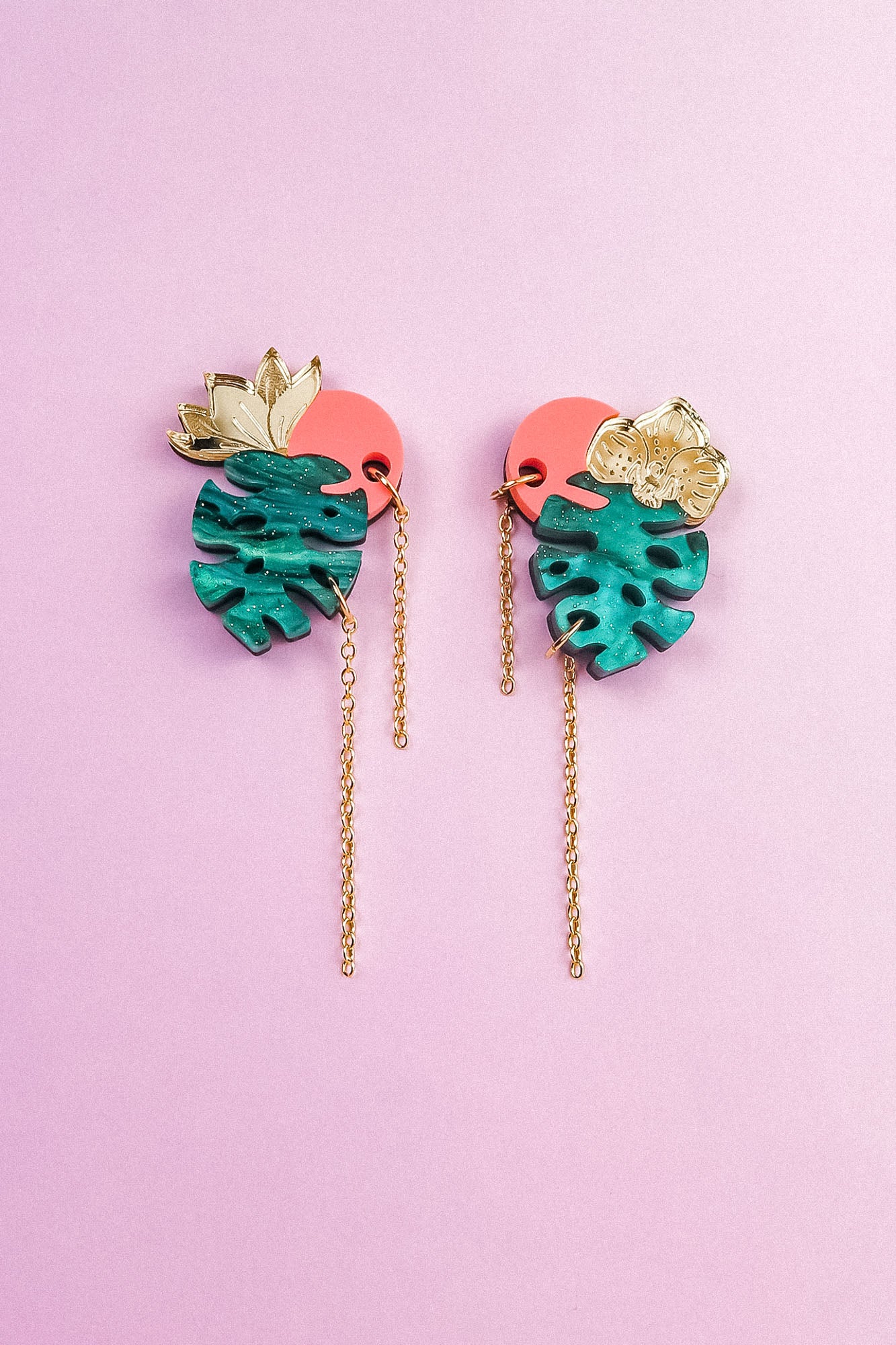A pair of colorful Tropicana Earrings with gold leaves, teal monstera-shaped pendants, pink circular accents, and long gold chains, featuring a handcrafted, tropical-inspired design on a light purple background.