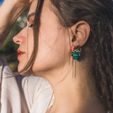 A woman with long brown hair and closed eyes tilts her head back, wearing a white top and handcrafted Tropicana Earrings featuring a tropical-inspired design with teal and gold details. Sunlight highlights her face and hair.