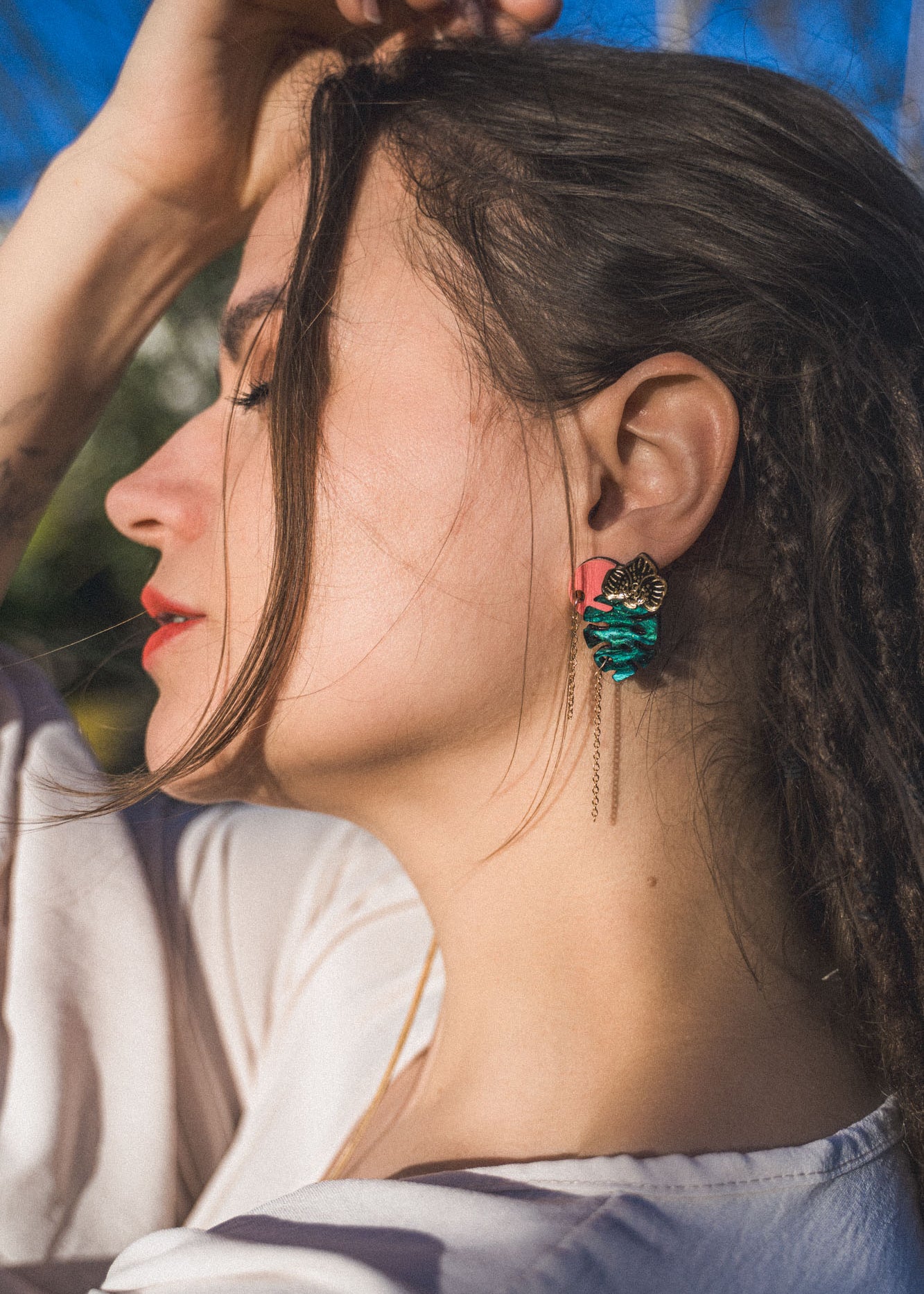 A woman with long brown hair and closed eyes tilts her head back, wearing a white top and handcrafted Tropicana Earrings featuring a tropical-inspired design with teal and gold details. Sunlight highlights her face and hair.