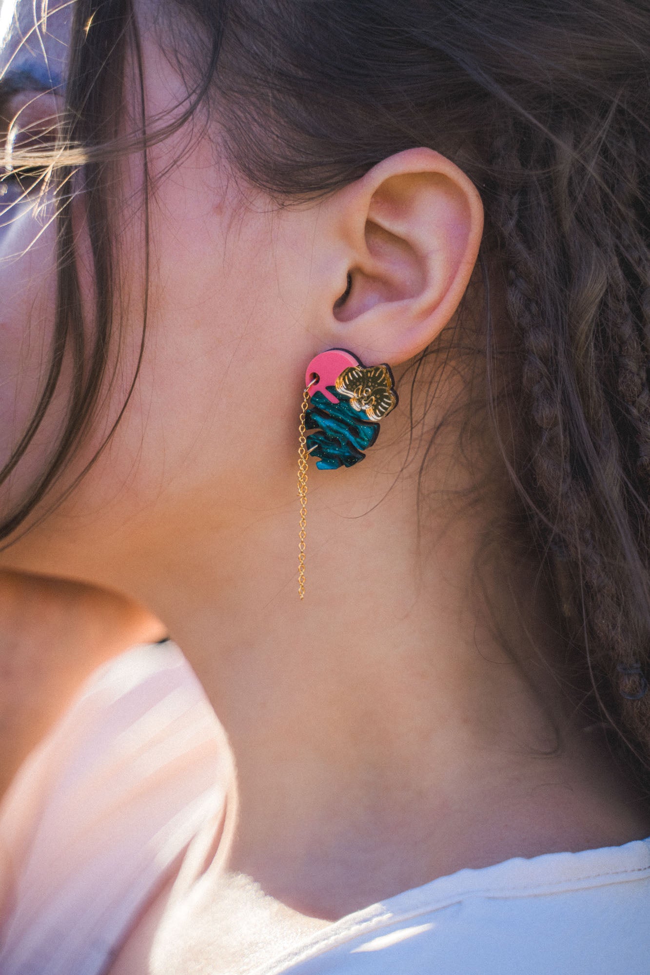 Close-up of a person’s ear wearing Tropicana Earrings—a tropical-inspired design with a gold tiger head, blue and pink details, and a delicate gold chain. The person’s hair is partially braided.