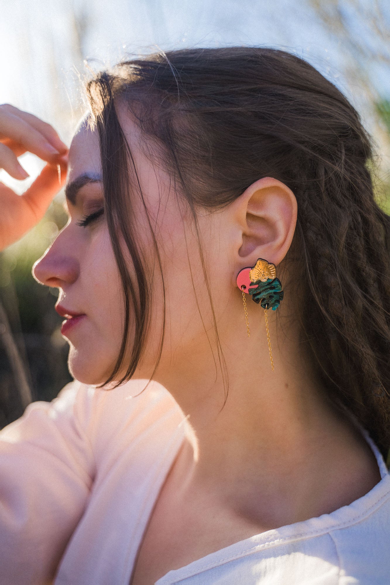 A woman with long brown hair in braids is shown in profile outdoors, wearing a pink top and handcrafted Tropicana Earrings with a tropical-inspired design shaped like leaves and a sun. Sunlight highlights her face and hair.
