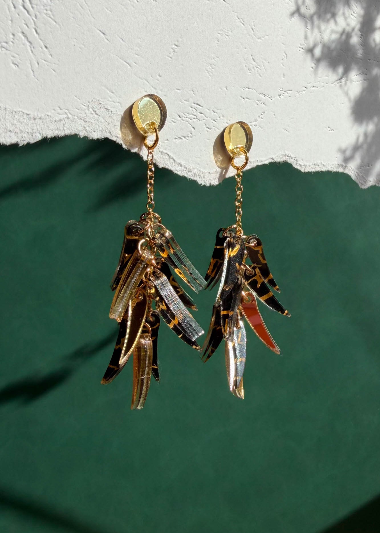 Featherlight gold and tortoiseshell dangle earrings with layered, leaf-like charms hang from a white textured surface above a green background. Light casts soft shadows on the wall.