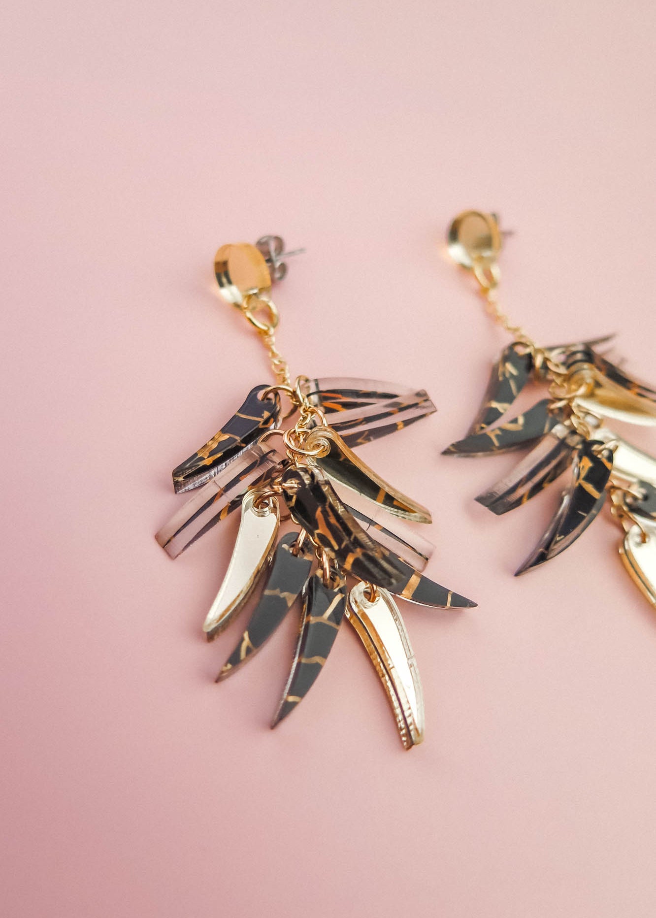 Gold and black marbled featherlight dangle earrings with multiple layered, leaf-shaped pendants arranged in a fan shape, displayed on a soft pink background.