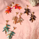 Colorful featherlight dangle earrings in four pairs—red-orange, brown-black, teal, and beige—feature acrylic tufts and are arranged on a pink surface with natural light, a beige rock, and wispy dried grass in the background.