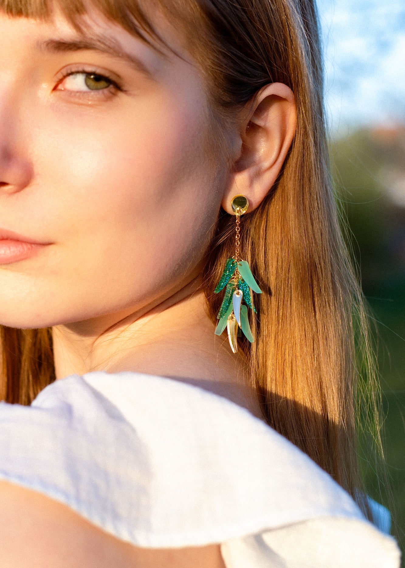 Close-up of a young woman with long light brown hair in a white top, showcasing featherlight dangle earrings shaped like green and gold leaves. She gazes slightly to the side, bathed in natural sunlight.