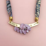 A handmade necklace featuring a pink and purple patterned fabric strap, a gold curved bar with small heart cutouts, and a cluster of marbled lavender decorative stones, displayed on a light pink background.