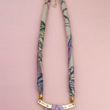 A handmade necklace with a fabric-wrapped band in muted colors and abstract patterns, featuring a gold bar centerpiece adorned with lilac stones, displayed on a light pink background—reminiscent of an elegant silk-print scarf.