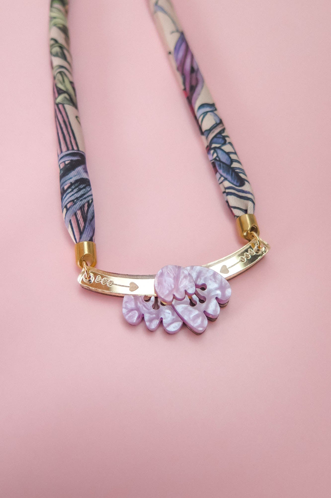 A handmade necklace with a patterned fabric band, gold metal bar, and three textured lilac stones, displayed on a pink background—perfect for pairing with an elegant silk-print scarf.