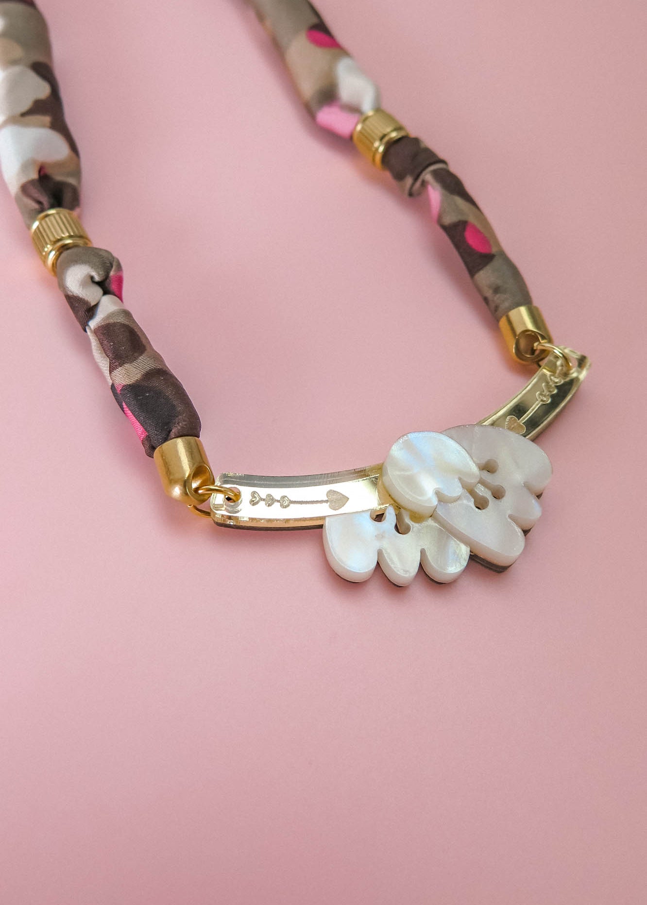 A close-up of a handmade necklace with a fabric-wrapped chain in brown, beige, and pink, gold details, and a white, carved mother-of-pearl pendant on a pink background.