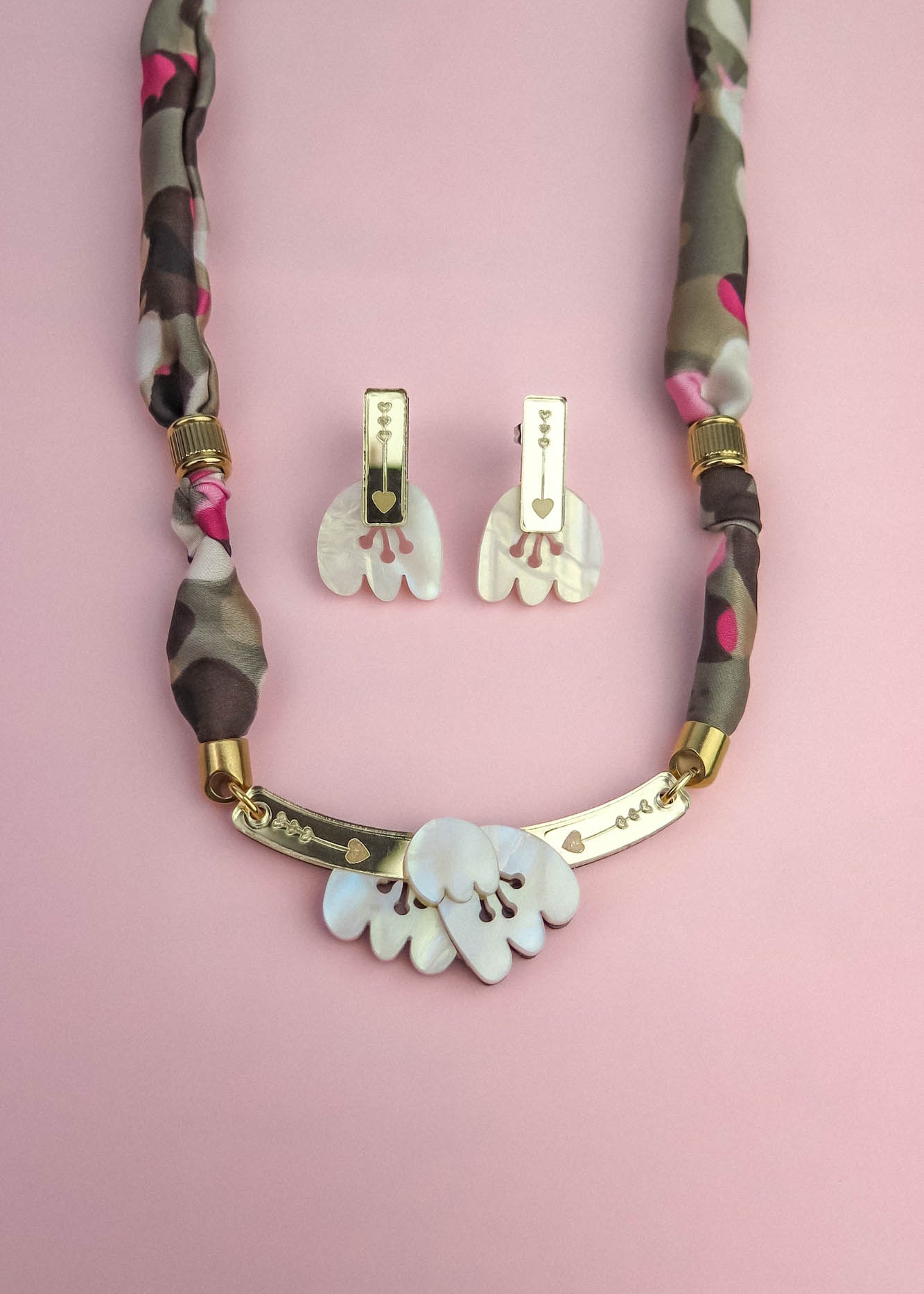 A handmade necklace with a fabric-wrapped chain and matching earrings, both featuring white, abstract, acrylic tulip-like pendants, displayed on a pink background.