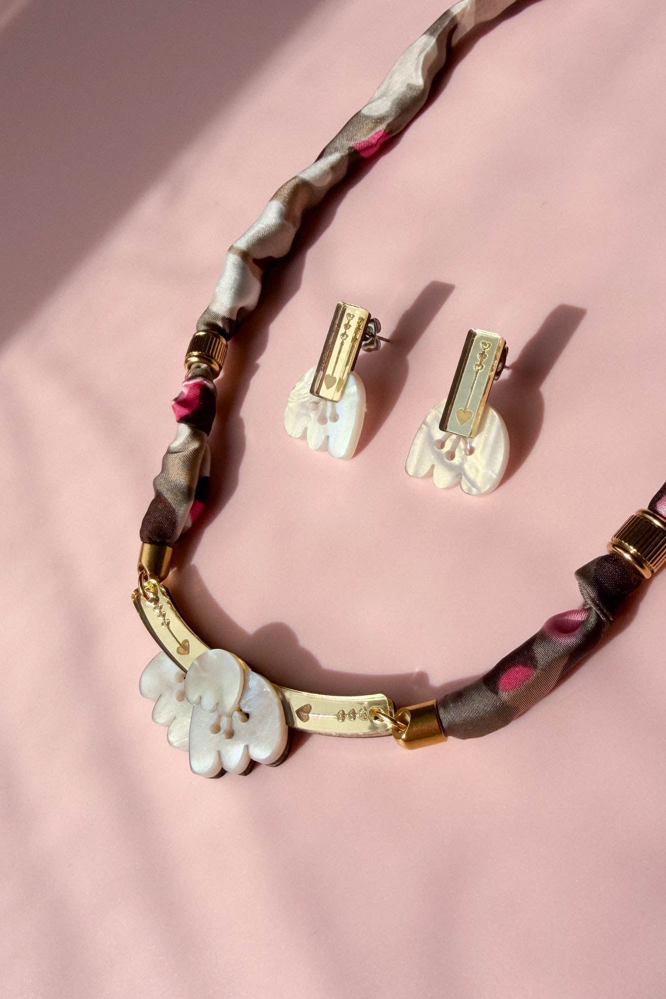 A handmade necklace and matching clip-on earrings with white, acrylic tulip-shaped pendants, gold accents, and a fabric-wrapped brown and pink chain, displayed on a soft pink background.