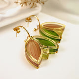 A pair of gold, leaf-shaped handcrafted earrings with translucent green and brown sections, displayed on a white surface with dried foliage in the background.