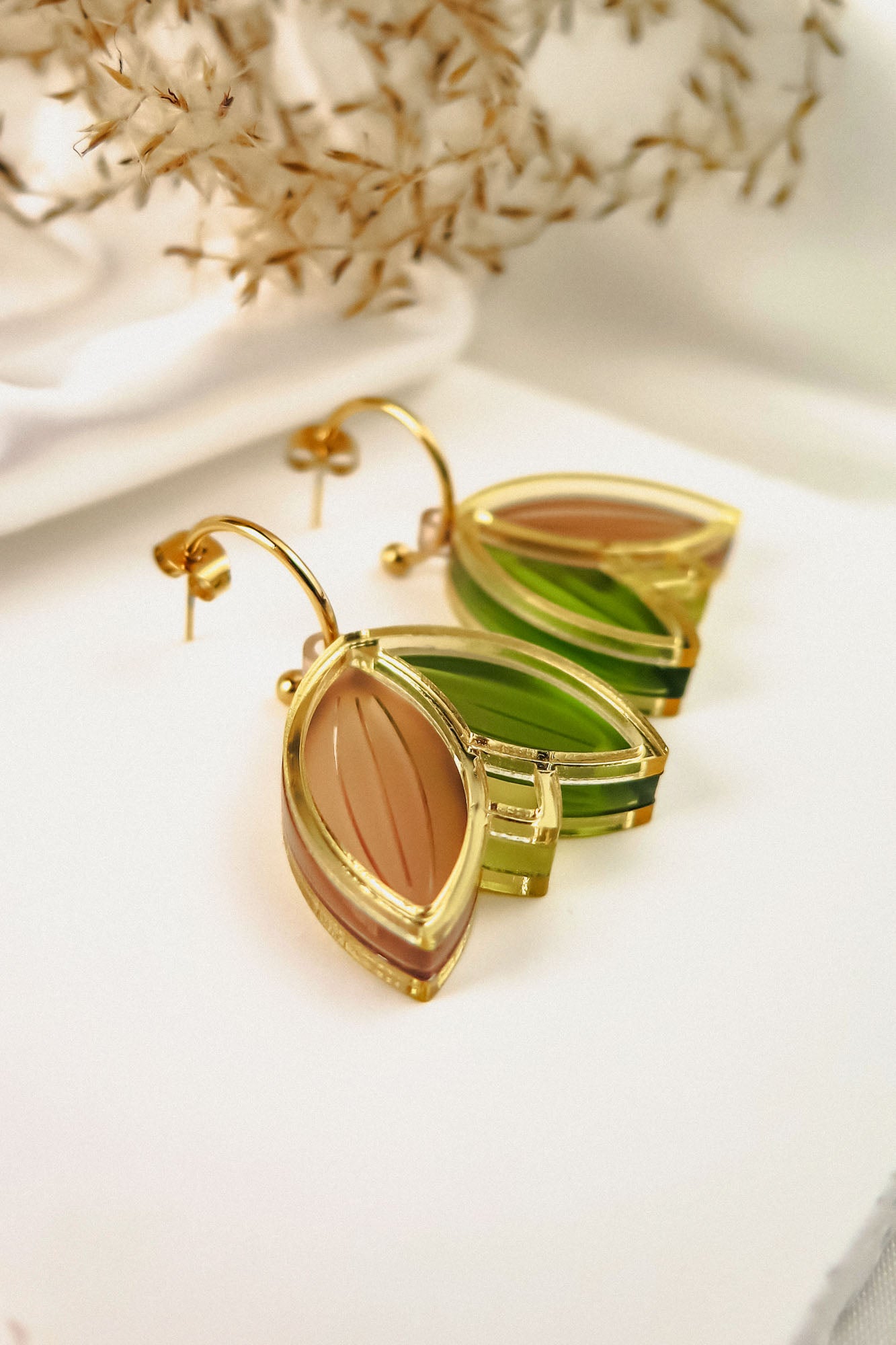 A pair of gold, leaf-shaped handcrafted earrings with translucent green and brown sections, displayed on a white surface with dried foliage in the background.