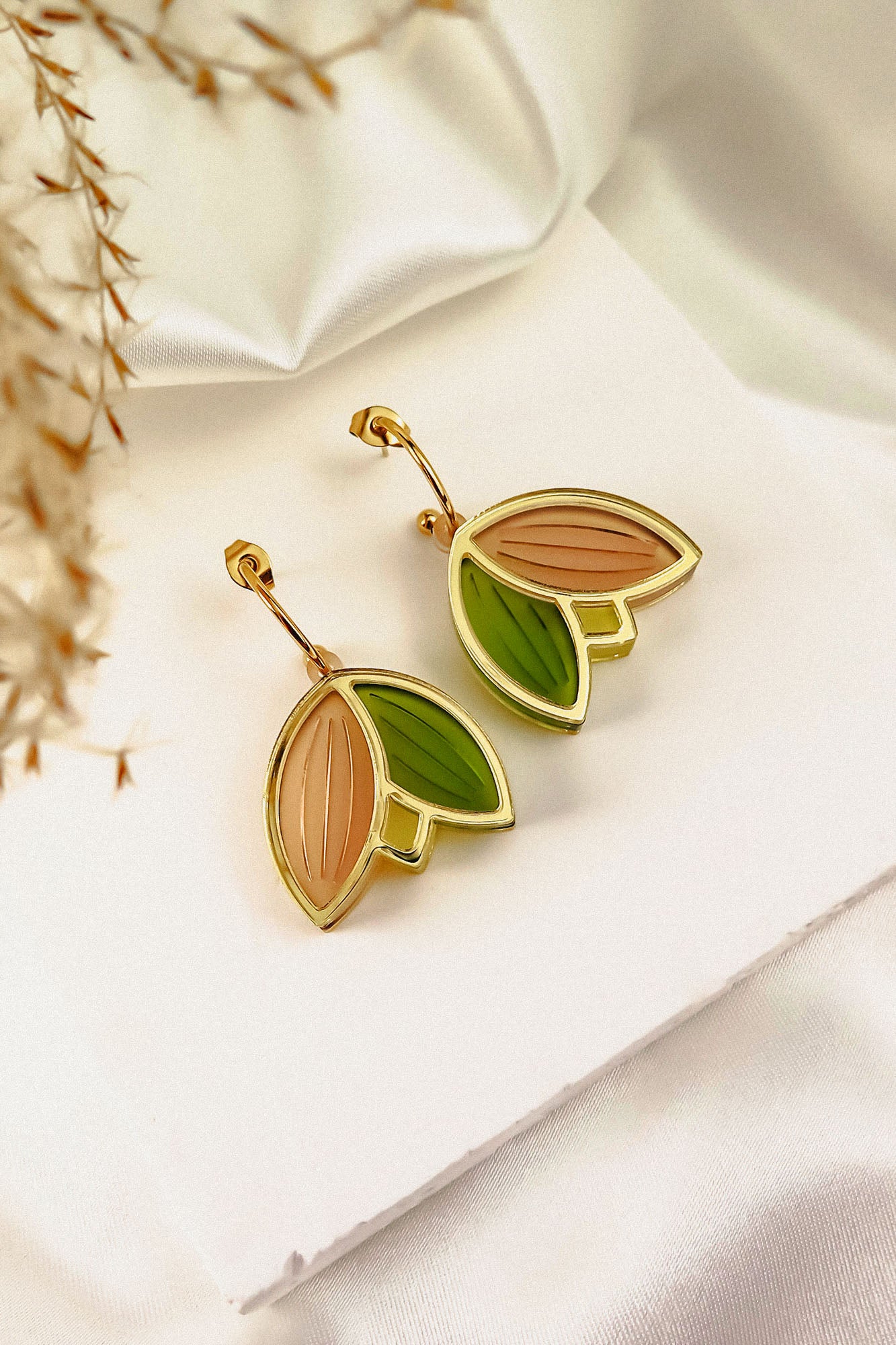 A pair of handcrafted gold outline earrings with leaf-shaped pendants in light pink and green enamel, displayed on a white card over satin fabric with dried grass in the corner. These lightweight earrings add a touch of elegance to any look.