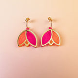A pair of Tulip Huggie Earrings with three petal-shaped sections—two orange and one bright pink—arranged in a floral design, showcasing artisan craftsmanship on a light peach gradient background.