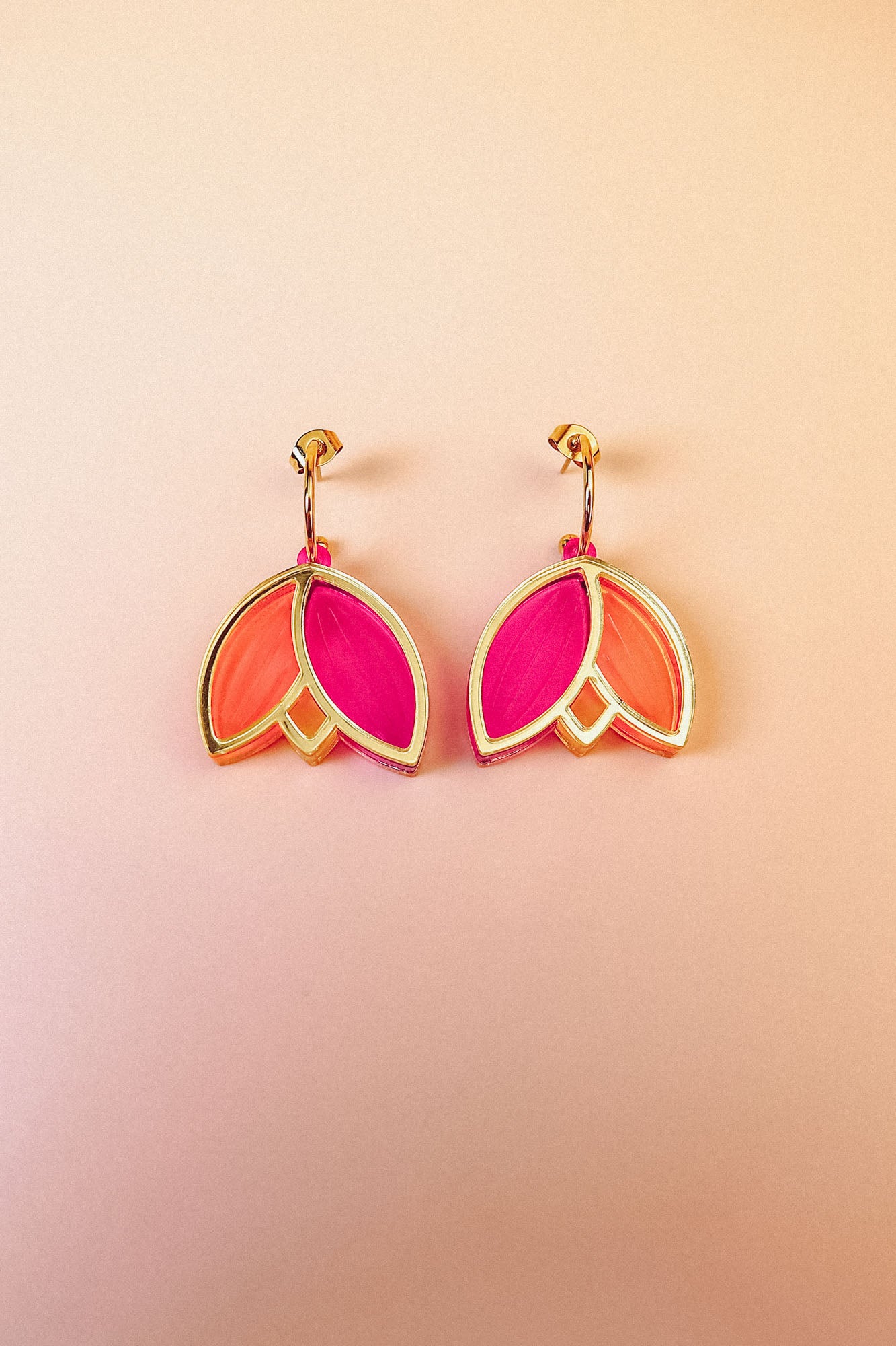 A pair of Tulip Huggie Earrings with three petal-shaped sections—two orange and one bright pink—arranged in a floral design, showcasing artisan craftsmanship on a light peach gradient background.