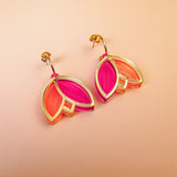 A pair of Tulip Huggie Earrings with artisan craftsmanship, featuring gold hoops and overlapping leaf-shaped charms in vibrant pink and orange colors, displayed on a peach-colored background.