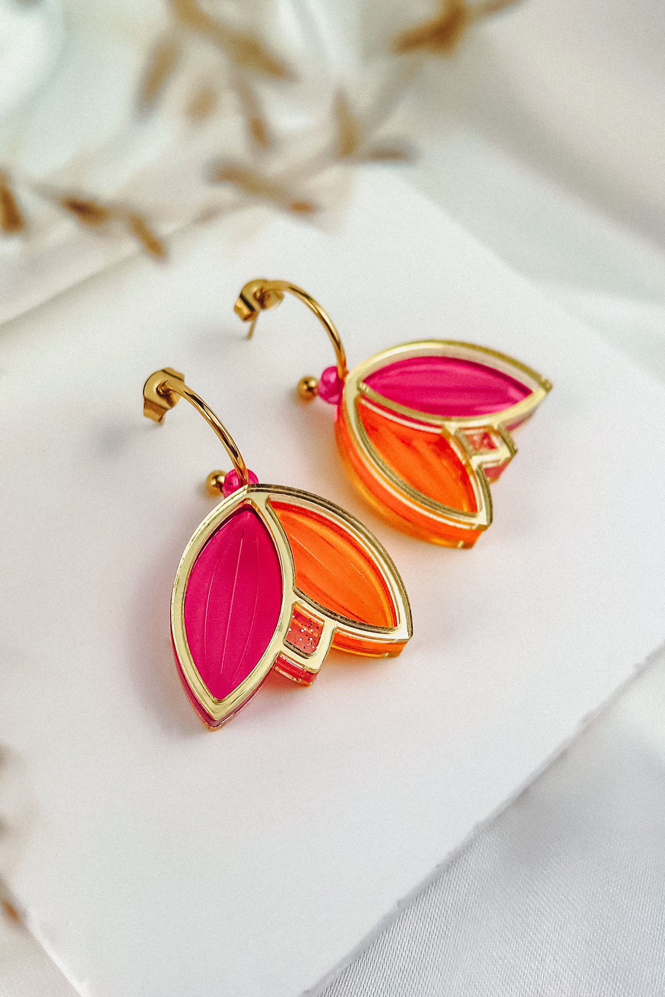 A pair of earrings with gold outlines, shaped like overlapping leaves in vibrant pink and orange colors, showcase artisan craftsmanship. Displayed on a white card with a soft, neutral background, these Tulip Huggie Earrings truly stand out.