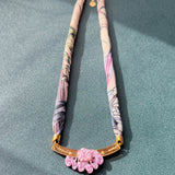 A handmade necklace with a fabric-wrapped band in pastel floral patterns, featuring a gold bar centerpiece adorned with pink flowers, and touches of acrylic tulip detail, displayed on a textured green surface.