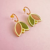 A pair of handcrafted gold metal Tulip Huggie Earrings with abstract leaf shapes in pink and green enamel, displayed on a light pink background. Lightweight earrings perfect for adding a pop of color to any look.