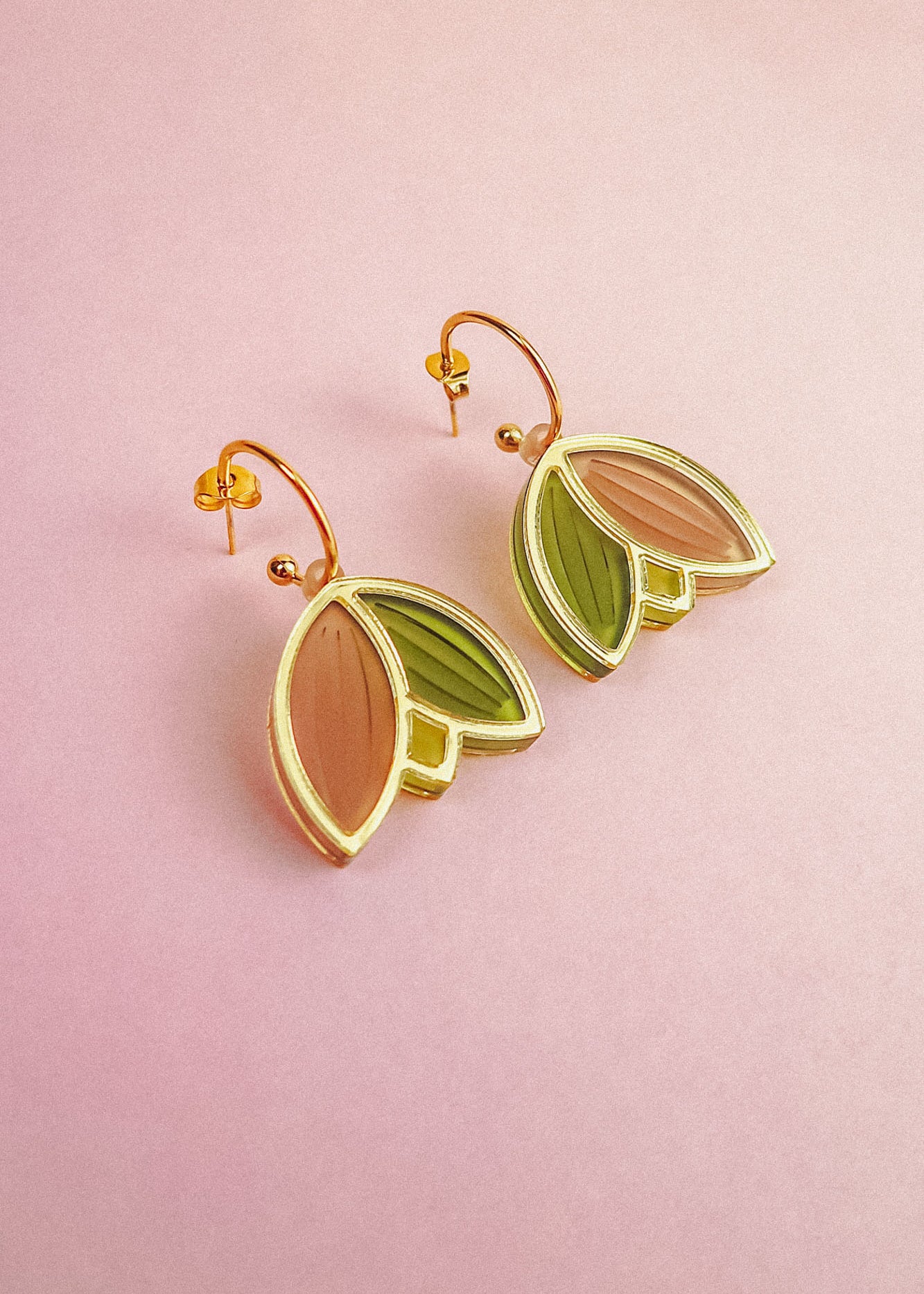 A pair of handcrafted gold metal Tulip Huggie Earrings with abstract leaf shapes in pink and green enamel, displayed on a light pink background. Lightweight earrings perfect for adding a pop of color to any look.