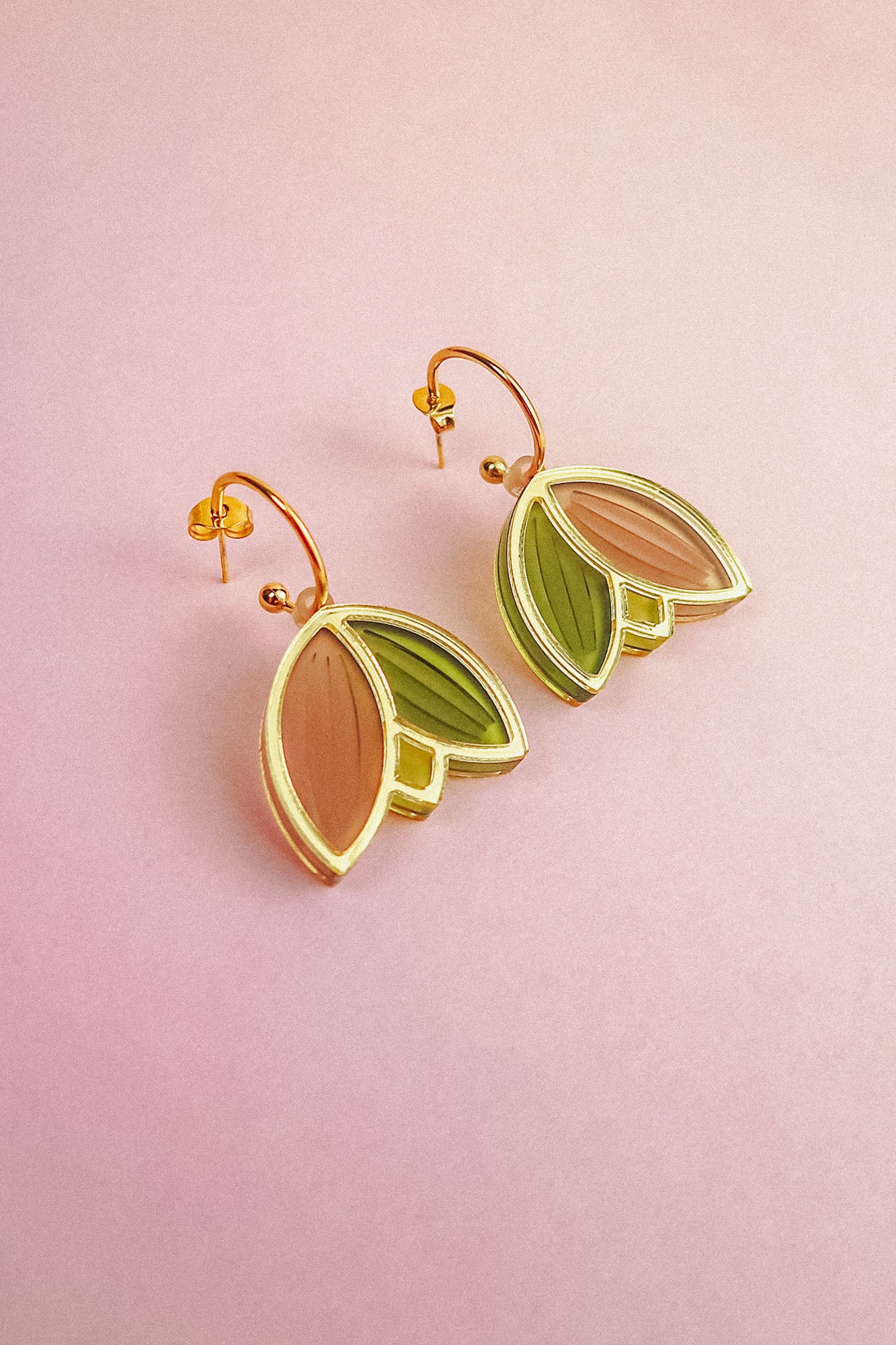 A pair of handcrafted gold metal Tulip Huggie Earrings with abstract leaf shapes in pink and green enamel, displayed on a light pink background. Lightweight earrings perfect for adding a pop of color to any look.