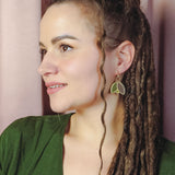 A woman with long, brown dreadlocks and a loose curl framing her face wears handcrafted leaf-shaped earrings and a green top, gazing to the left against a backdrop of soft pink curtains.