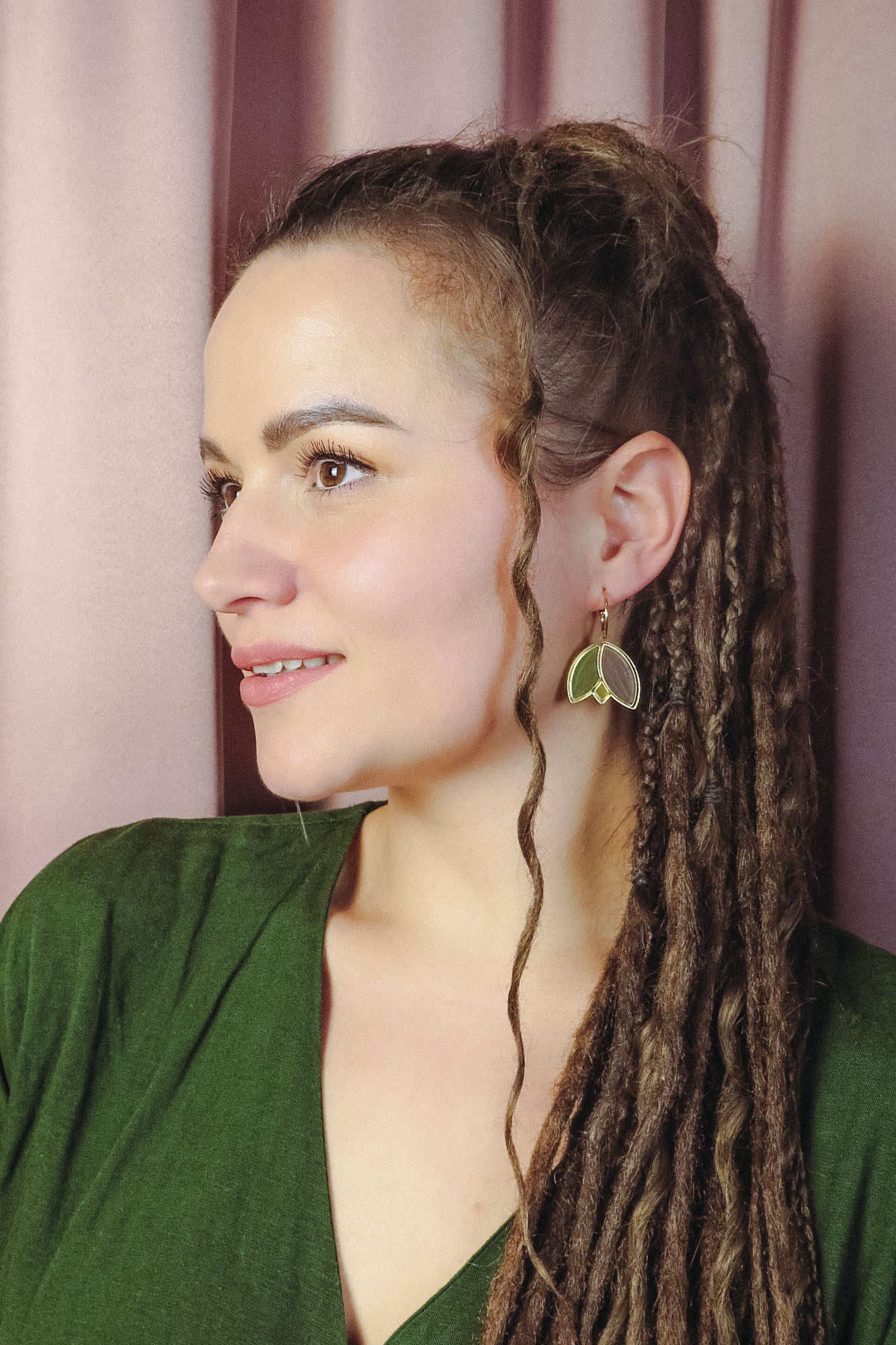 A woman with long, brown dreadlocks and a loose curl framing her face wears handcrafted leaf-shaped earrings and a green top, gazing to the left against a backdrop of soft pink curtains.