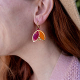 A person with red hair wearing a tan hat and pink sweater is shown in profile, displaying Tulip Huggie Earrings crafted with artisan craftsmanship and featuring gold, pink, and orange enamel sections.
