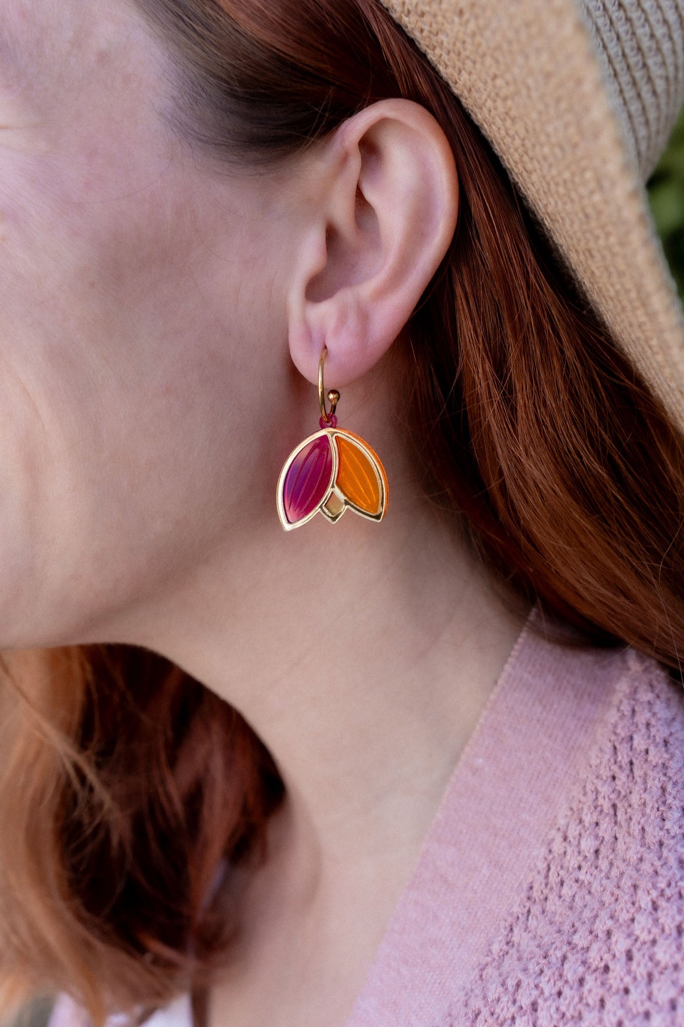 A person with red hair wearing a tan hat and pink sweater is shown in profile, displaying Tulip Huggie Earrings crafted with artisan craftsmanship and featuring gold, pink, and orange enamel sections.