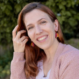 A woman with long red hair and Tulip Huggie Earrings smiles outdoors, wearing a light pink sweater over a white top. Green foliage is blurred in the background, highlighting the earrings vibrant colors and artisan craftsmanship.