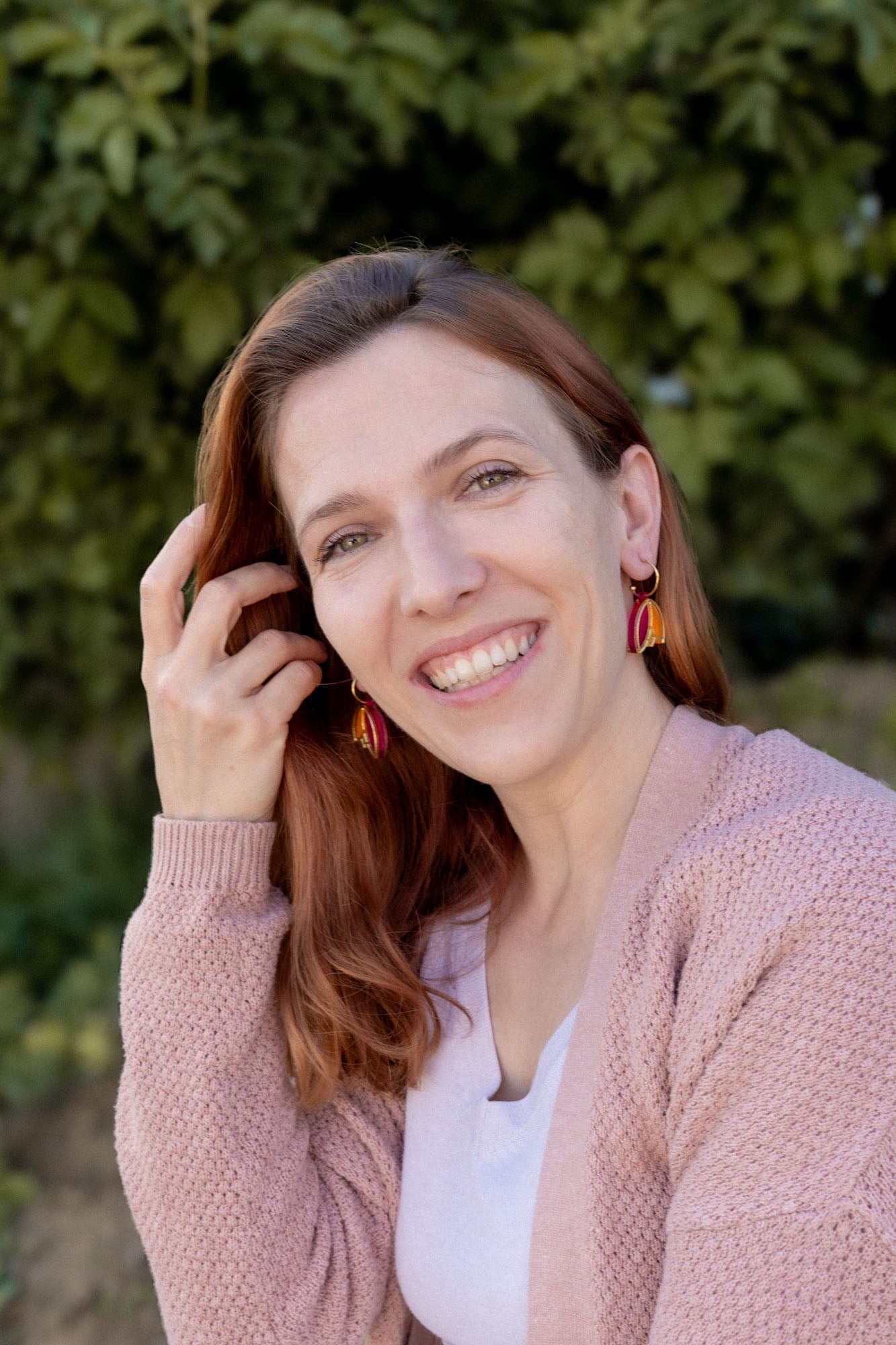 A woman with long red hair and Tulip Huggie Earrings smiles outdoors, wearing a light pink sweater over a white top. Green foliage is blurred in the background, highlighting the earrings vibrant colors and artisan craftsmanship.