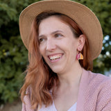 A woman with wavy reddish hair, wearing a straw hat, pink cardigan, white top, and Tulip Huggie Earrings featuring vibrant colors, smiles while standing outdoors in front of green foliage.