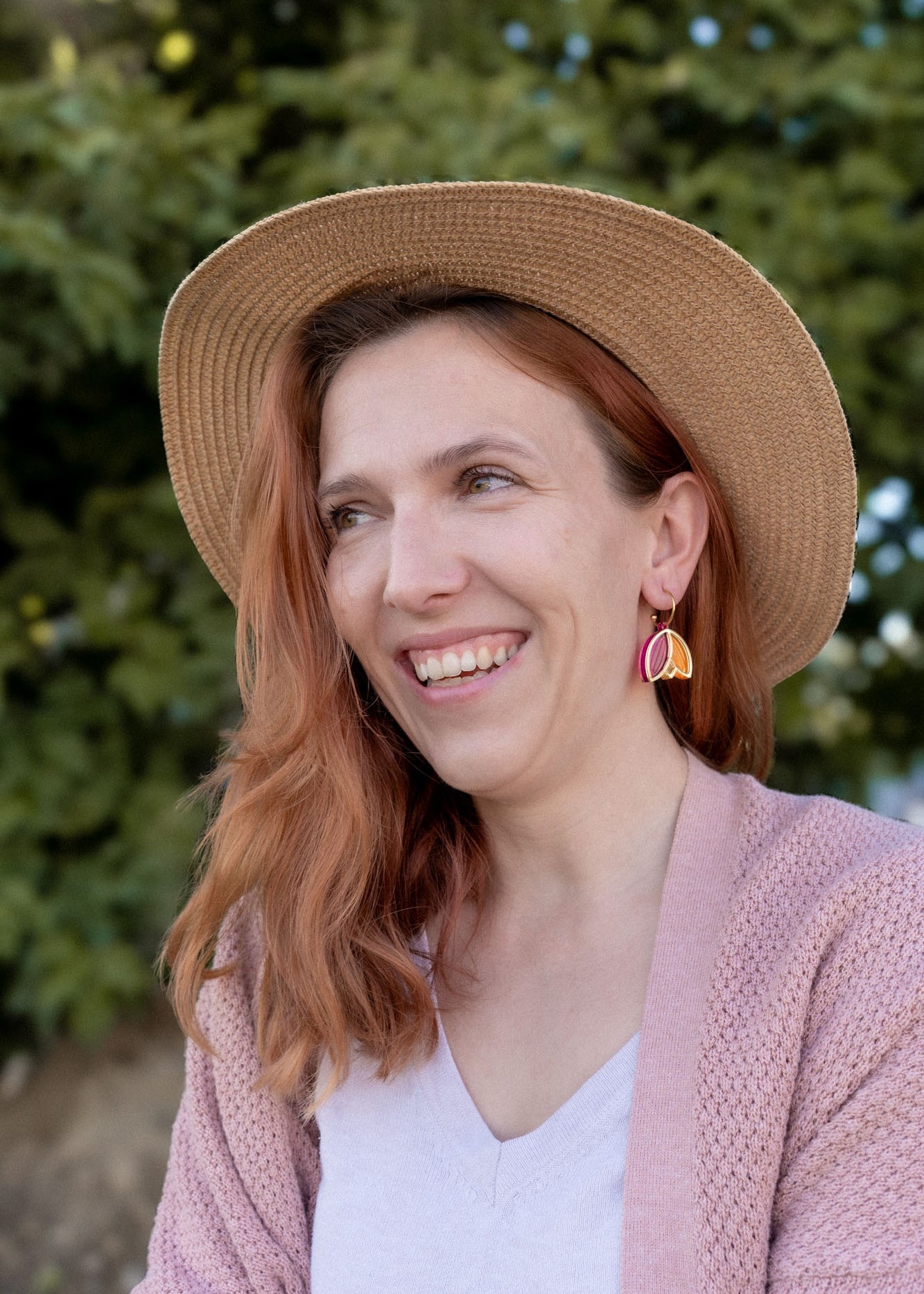 A woman with wavy reddish hair, wearing a straw hat, pink cardigan, white top, and Tulip Huggie Earrings featuring vibrant colors, smiles while standing outdoors in front of green foliage.