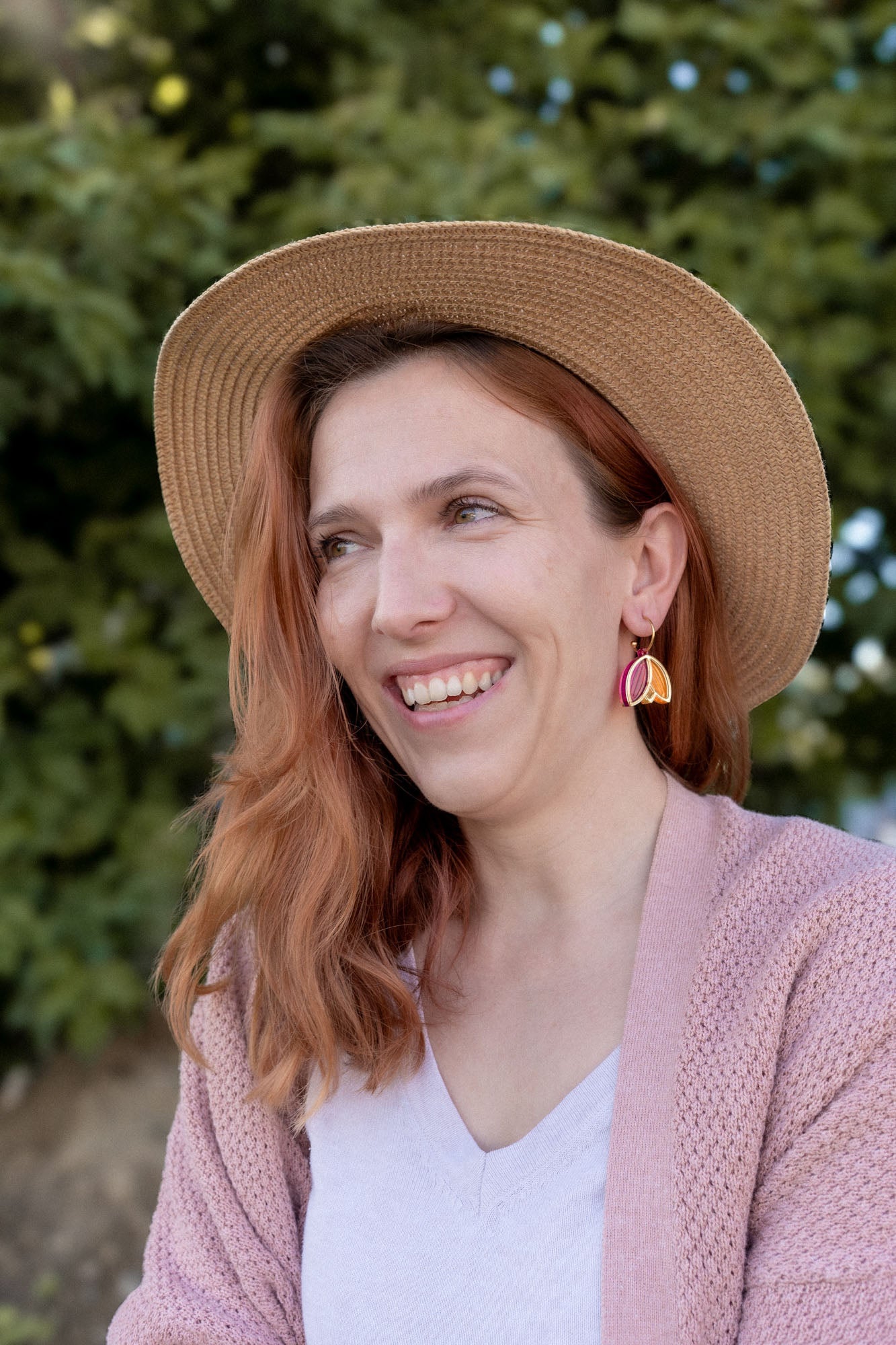 A woman with wavy reddish hair, wearing a straw hat, pink cardigan, white top, and Tulip Huggie Earrings featuring vibrant colors, smiles while standing outdoors in front of green foliage.