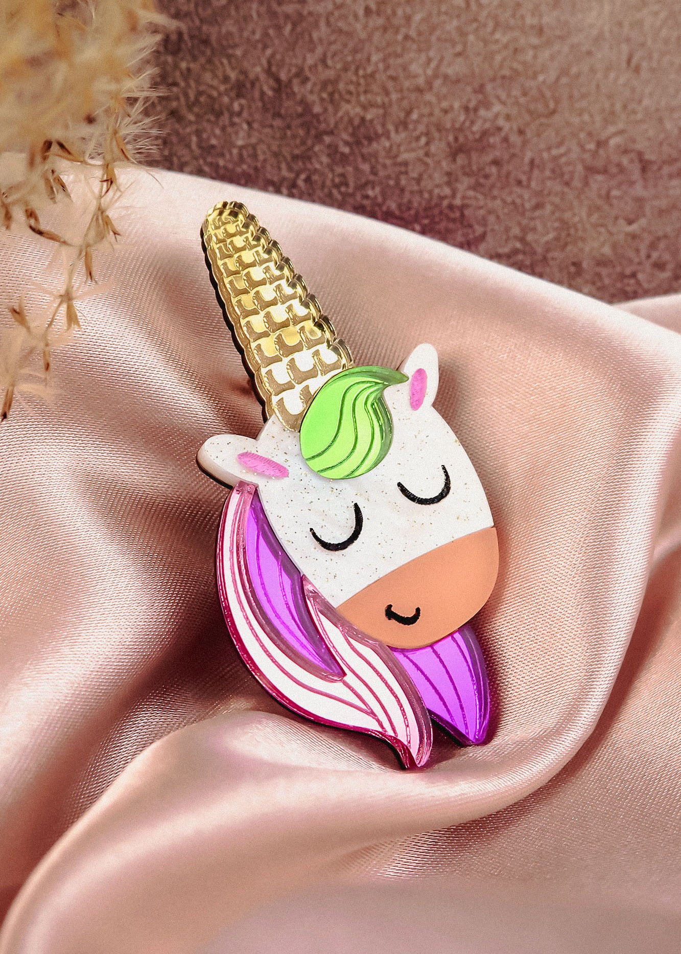 A cute handcrafted UniCorn Brooch with a gold horn, green mane, pink-and-purple hair, and closed eyes rests on pink satin fabric—a playful accessory for any outfit.