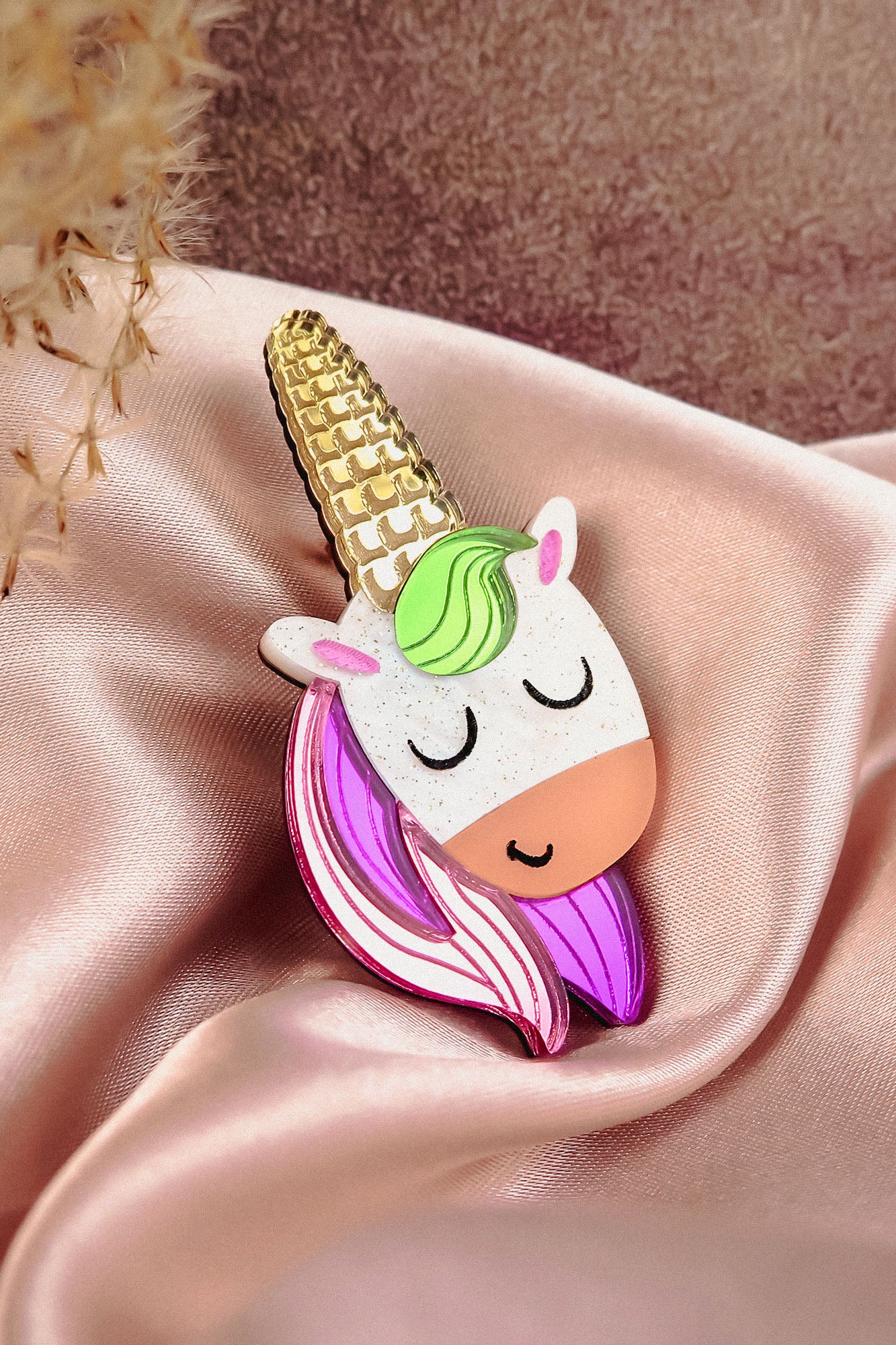 A cute handcrafted UniCorn Brooch with a gold horn, green mane, pink-and-purple hair, and closed eyes rests on pink satin fabric—a playful accessory for any outfit.