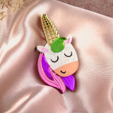 A playful accessory, this handcrafted UniCorn Brooch features a colorful unicorn with closed eyes, a gold horn, and pink and purple mane, resting on shiny pale pink satin fabric with dried flowers in the corner.