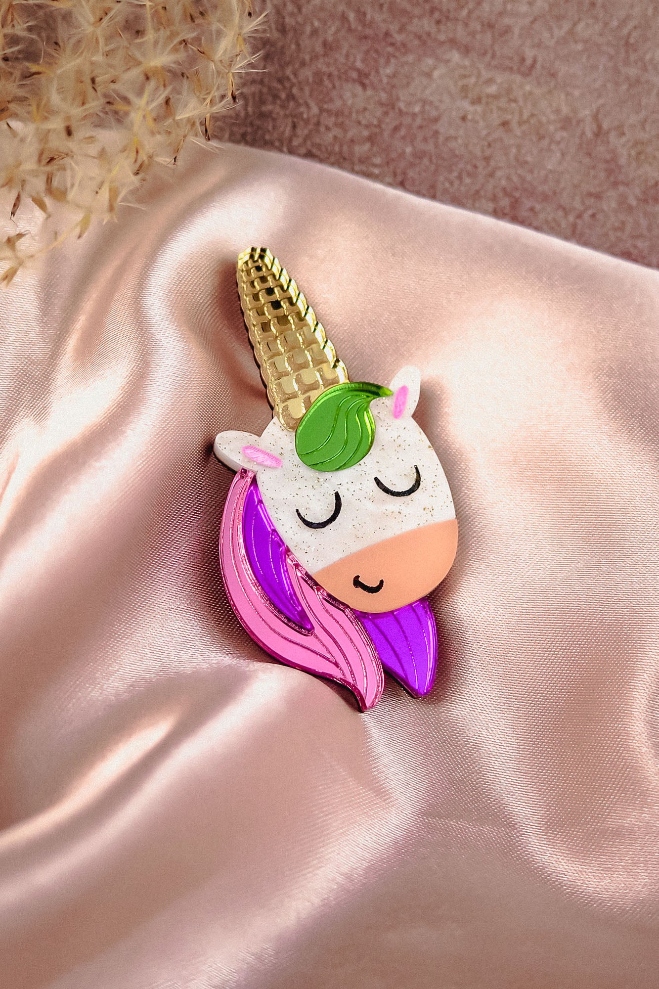 A playful accessory, this handcrafted UniCorn Brooch features a colorful unicorn with closed eyes, a gold horn, and pink and purple mane, resting on shiny pale pink satin fabric with dried flowers in the corner.