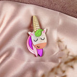 A handcrafted UniCorn Brooch with a gold horn, green and pink mane, and closed eyes rests on soft, shiny pink fabric. A blurred plant is visible in the foreground, highlighting this playful accessory’s charm.