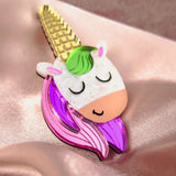 A colorful handcrafted UniCorn Brooch shaped like a unicorn’s head with a golden horn, green mane, closed eyes, and pink and purple details rests on shiny light pink satin fabric—a playful accessory for any collection.