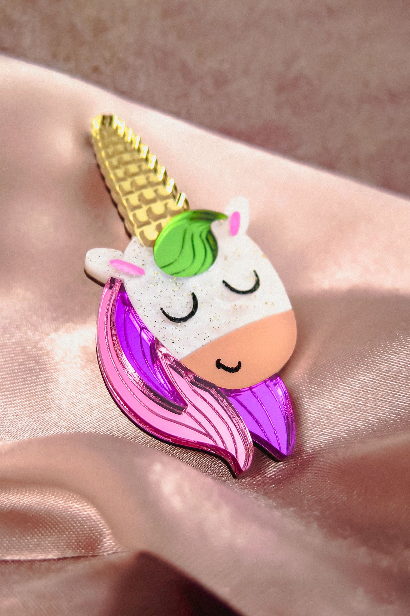 A colorful handcrafted UniCorn Brooch shaped like a unicorn’s head with a golden horn, green mane, closed eyes, and pink and purple details rests on shiny light pink satin fabric—a playful accessory for any collection.