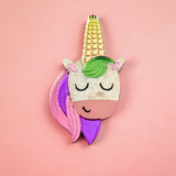 A playful accessory, this handcrafted UniCorn Brooch features a cute unicorn face with closed eyes, a golden horn, green mane, and pink and purple hair on a pastel pink background.