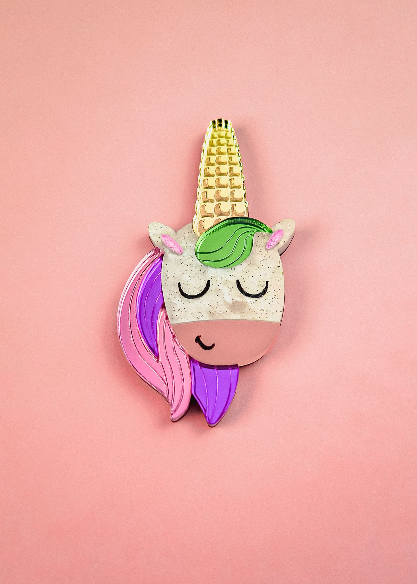 A playful accessory, this handcrafted UniCorn Brooch features a cute unicorn face with closed eyes, a golden horn, green mane, and pink and purple hair on a pastel pink background.